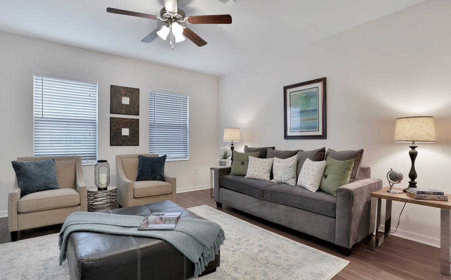 Spacious living room with grey sofa with light color pillows, brown leather ottoman with blue blanket on it, and two light color chairs on the back wall.