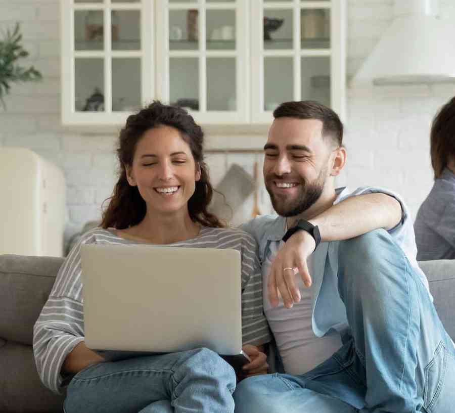 Couple sitting on a gray couch looking at a laptop, while two children play in the room.