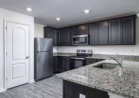 Open-concept kitchen with installed appliances, upper-wood cabinets and large granite countertops