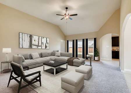Staged spacious living room with neutral palette, two tone paint, beige carpet, ceiling fan, three windows to backyard with drapes and blinds, sectional sofa, chairs, area rug, coffee and end tables and lamp with artwork and archways to dining and kitchen.