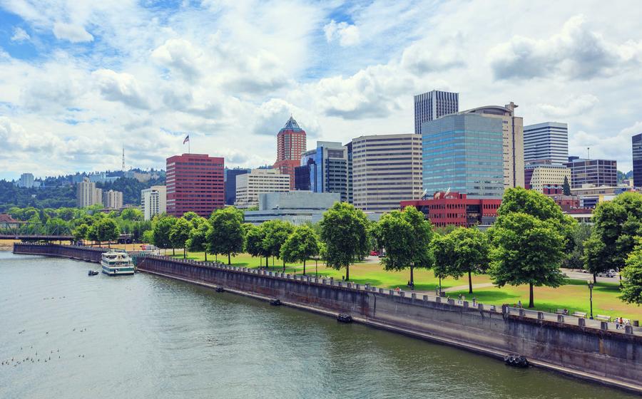 Portland, Oregon skyline taken from the river showing a docked boat, trees and grass on the bank, and office buildings