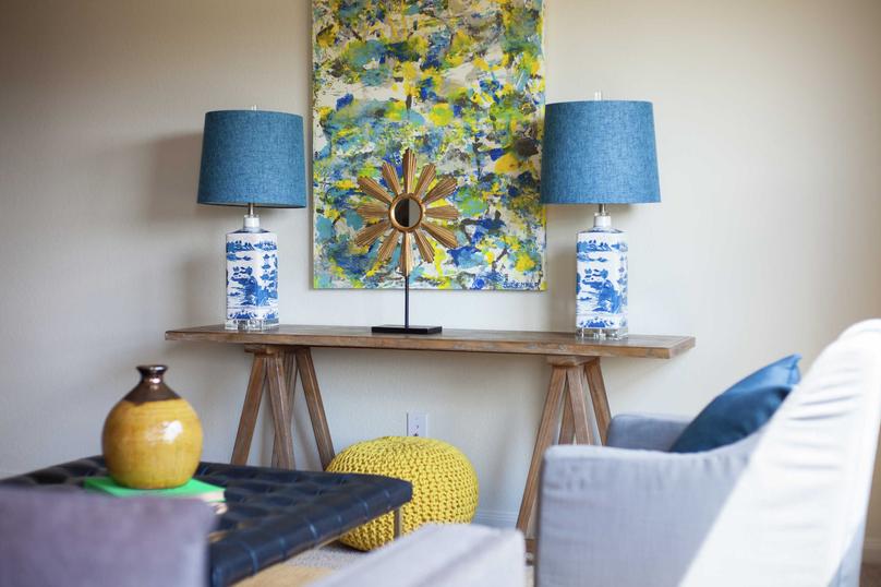 Staged living room with two decorative lamps with blue shades on a wooden table, painting hanging on the wall and a yellow vase on a navy coffee table.