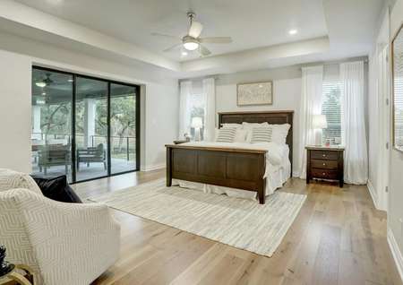 Staged master bedroom with tall ceilings, white walls, light wood flooring and back yard views.