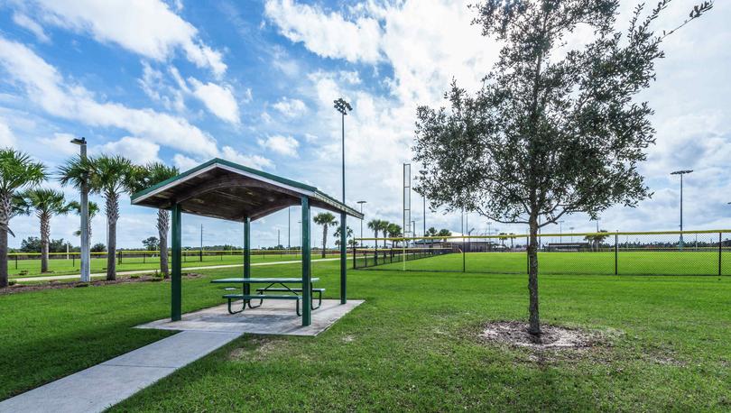 A covered bench area that is surrounded by grass with a gated sports field that has light in the Poinciana community.