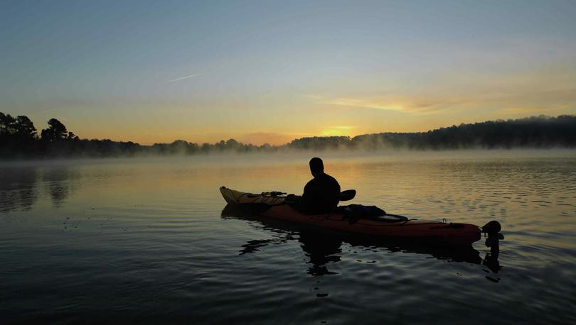 Kayaker silhouette paddling across water with treeline horizon in the background