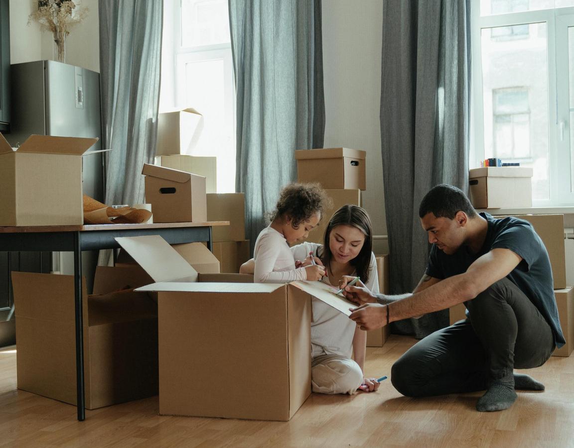 A young family sits on the floor drawing on a box and is surrounded by moving boxes.