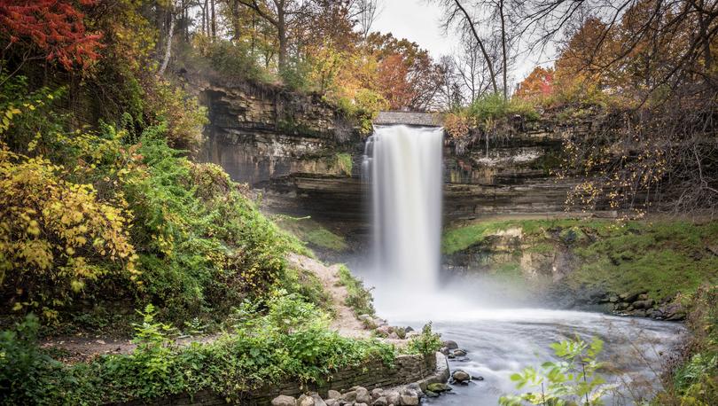 Minneapolis, Minnesota Minnehaha Falls with water pouring over the cliff into the stream below, rocks along the bank, and lush green plants