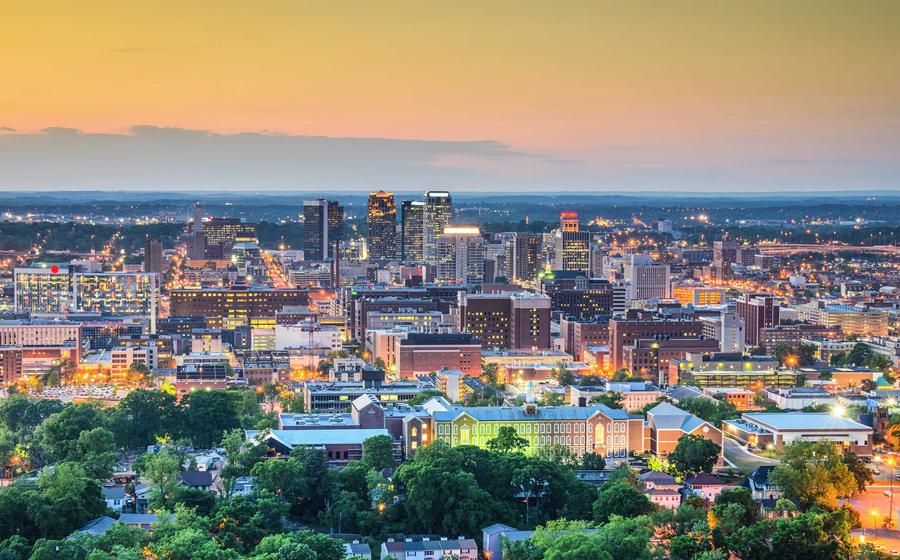 Birmingham, Alabama downtown at dusk with tree-covered residential neighborhoods, lit windows in apartments, and skyscrapers in the distance