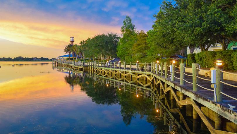 The Villages, Florida wooden boardwalk at dusk with lights on, lined with green trees, and calm lake waters