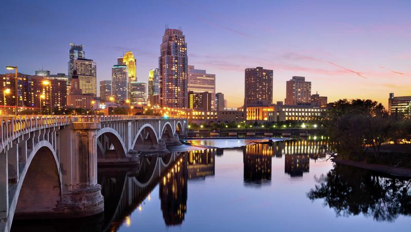 Minneapolis, Minnesota Skyline showing Arch Bridge spanning across the water, windows lit in skyscrapers, and the sun setting on the horizon