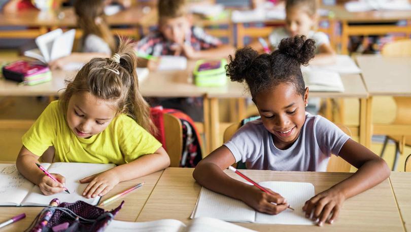 School room full of children with African American girl wearing light purple t-shirt writing on paper and Caucasian girl with yellow shirt also writing sitting in the front