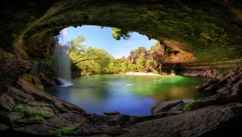 Austin, Texas Hamilton Pool waterfall recreational area with stone cave, water falling over the cliff, and trees off in the distance