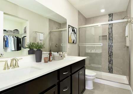 Staged master bathroom with two sinks and quartz countertop on vanity, dark brown cabinets, a shower/tub combo with glass door and decorative tile accents, view into attached walk-in closet.