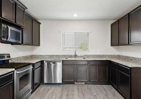 Photo of open kitchen with dark brown cabinets, gray speckled counters, stainless appliances and plank flooring.