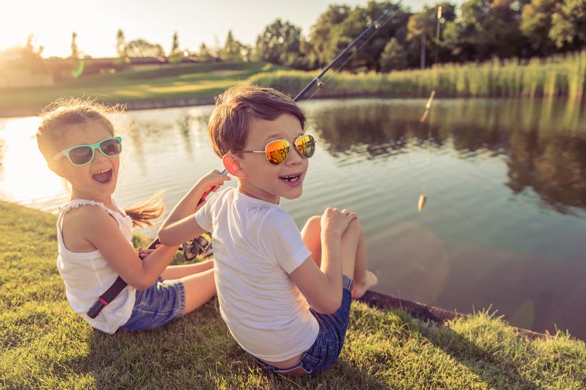Funny stylish little boy and girl in sun glasses are looking at camera and smiling while catching fish in the pond using a fishing rod, sitting on the ground