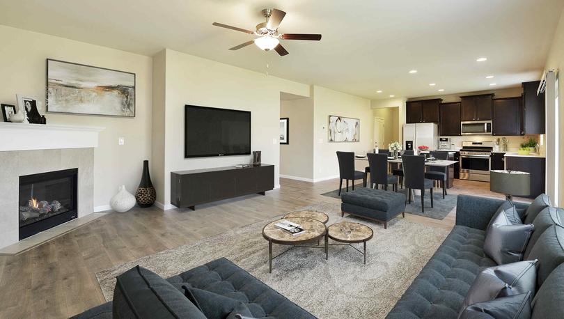 Decorated living room with plank flooring, a corner fireplace, ceiling fan and open to kitchen with dark cabinets and stainless appliances.