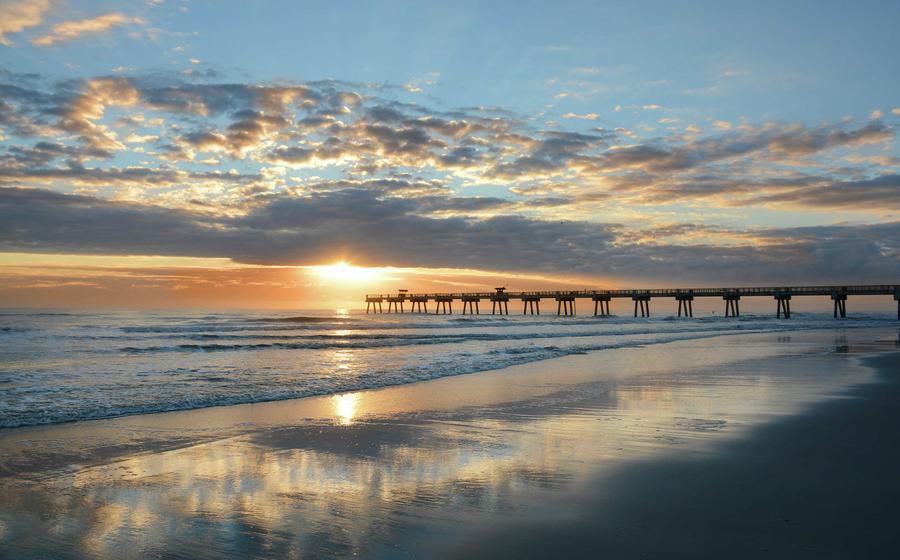 Jacksonville, Florida beach sunrise with pier extended far into the ocean, rolling waves, and sunlight reflecting off the wet sand