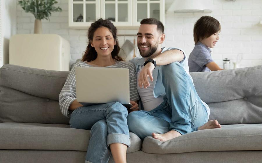 Couple sitting on a gray couch looking at a laptop, while two children play in the room.