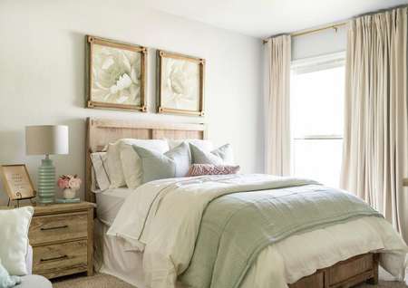 Staged secondary bedroom with artwork above the bed, a nightstand and large window with curtains. 