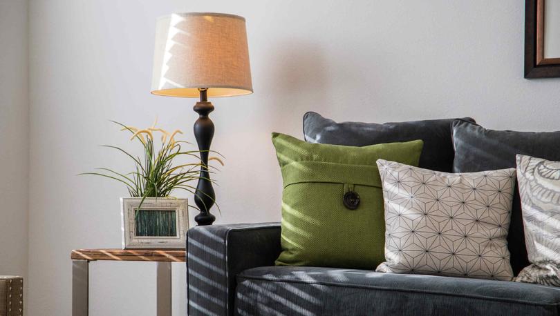 Model home with dark blue sofa that has green and white pillows, framed picture hanging on the wall and lamp with beige shade in the corner. 