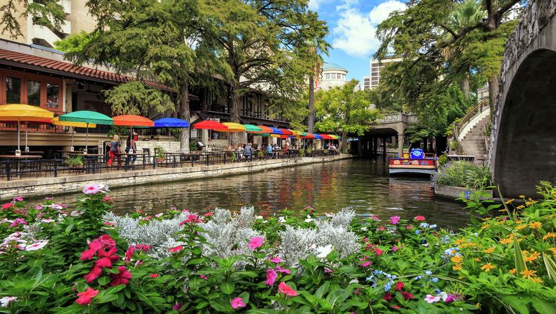 San Antonio, Texas Riverwalk showing blooming pink, white, and yellow flowers, multi-color sunshades and tables lining the river, and overgrown trees