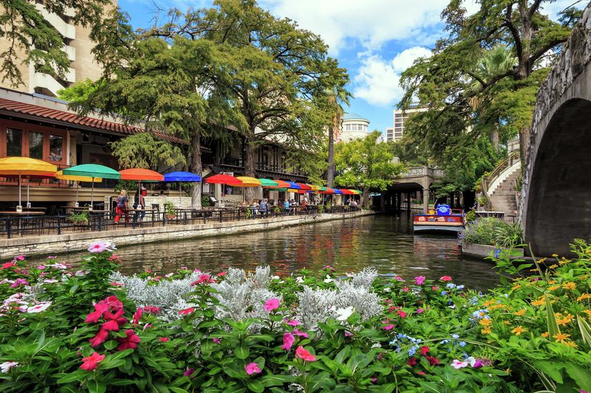 San Antonio, Texas Riverwalk showing blooming pink, white, and yellow flowers, multi-color sunshades and tables lining the river, and overgrown trees