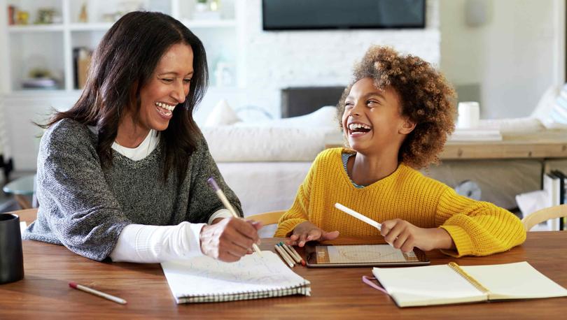 Happy mom and child work on homework at table with sofa and tv in background.