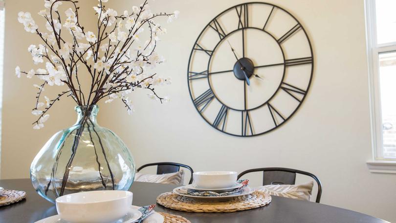 Maple model home dining nook staged with wooden table with placemats, white bowels, and glass ornament with flowers plus large wall clock in the back