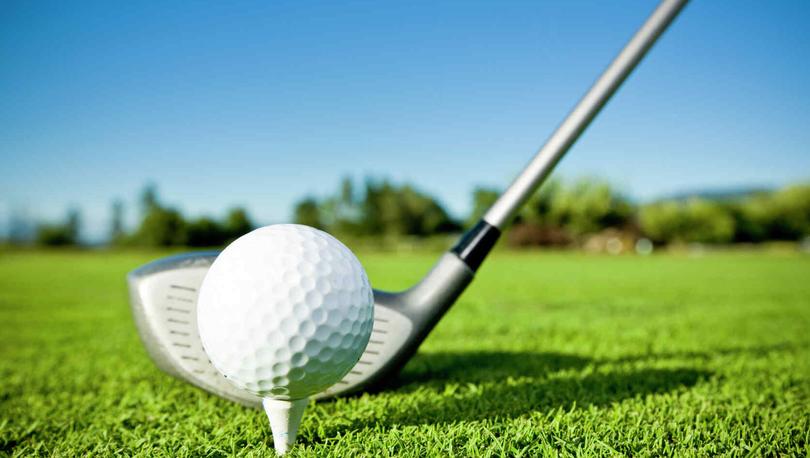 Closeup of golf club lined up to hit a white golf ball on white tee, green grass, trees in background, blue sky.