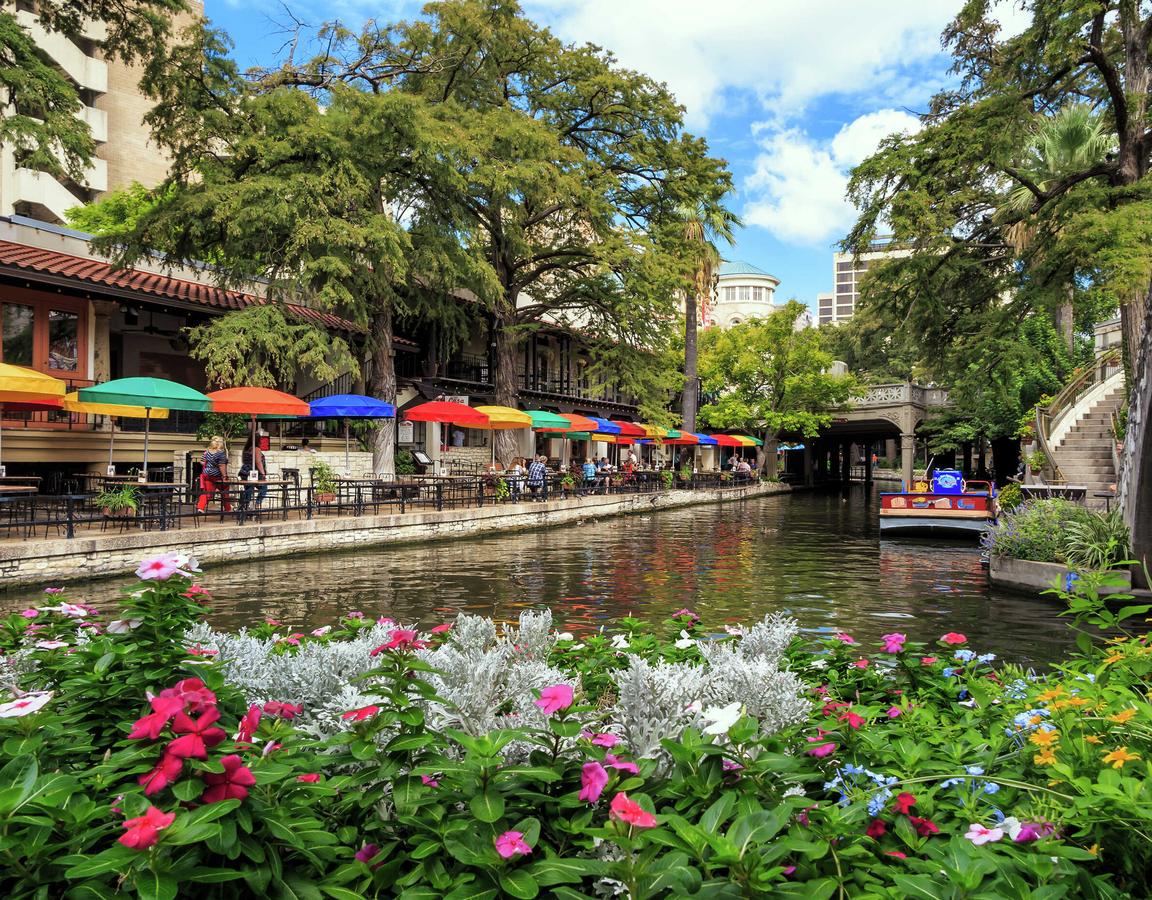 San Antonio, Texas Riverwalk showing blooming pink, white, and yellow flowers, multi-color sunshades and tables lining the river, and overgrown trees