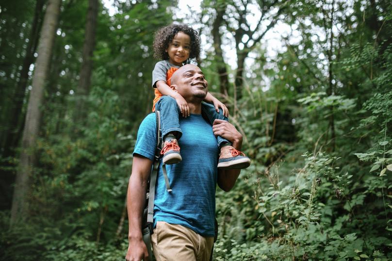 A smiling dad holds his boy on his shoulders while hiking in the forest.