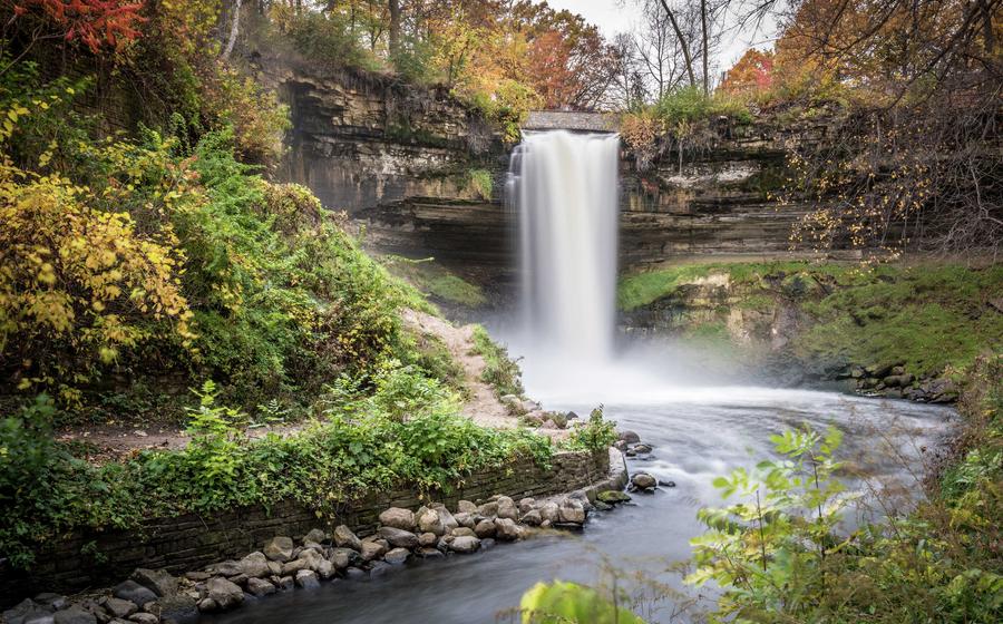 Minneapolis, Minnesota Minnehaha Falls with water pouring over the cliff into the stream below, rocks along the bank, and lush green plants