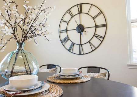 Wooden table with placemats, white bowls and glass ornament with flowers plus large wall clock in the back.