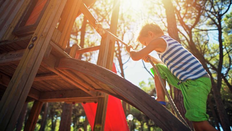 Boy with green shorts and striped sleeveless shirt climbing on wooden playground equipment