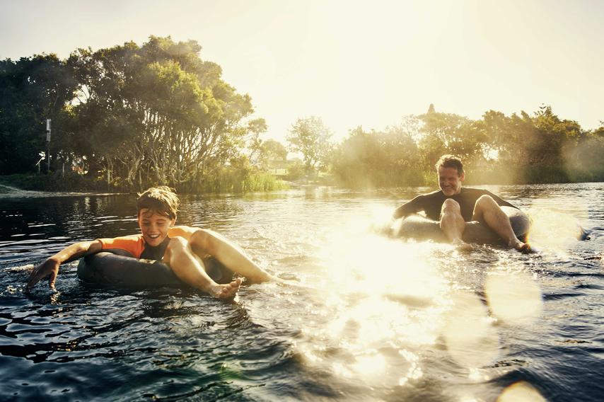 Father and son drifting down river on black intertubes with trees in the background