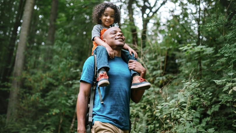 A smiling dad holds his boy on his shoulders while hiking in the forest.