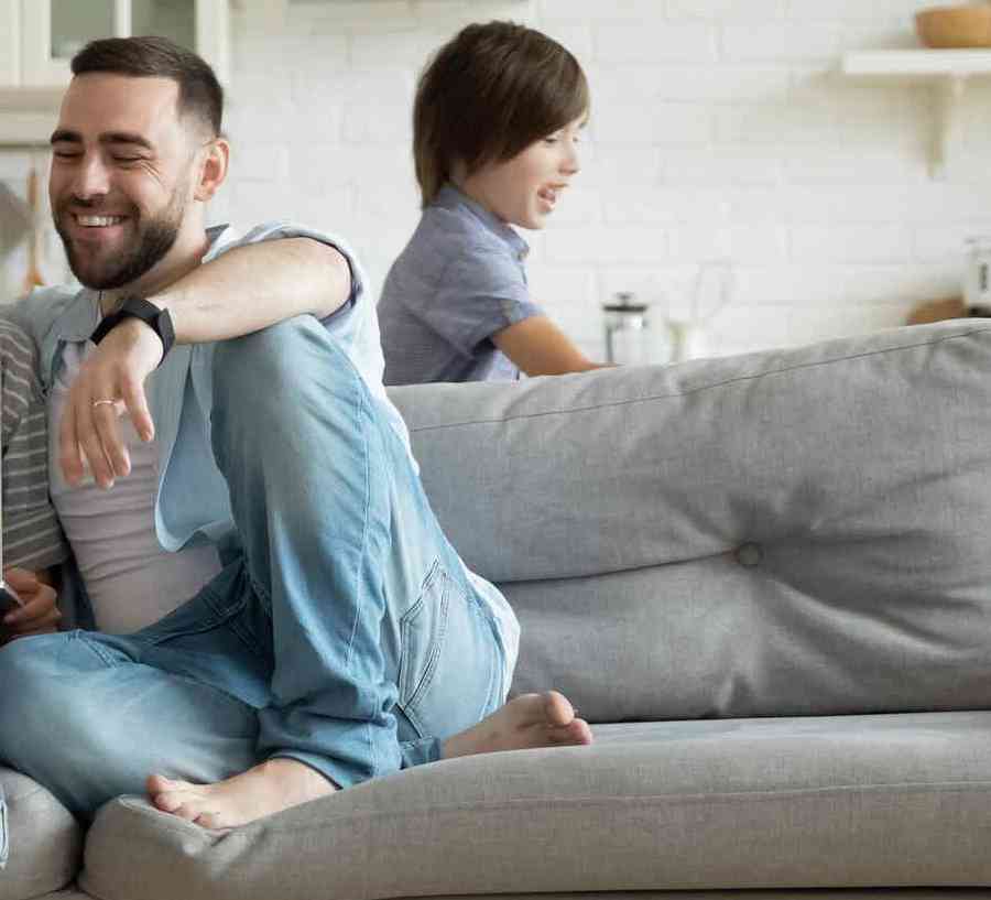 Couple sitting on a gray couch looking at a laptop, while two children play in the room.